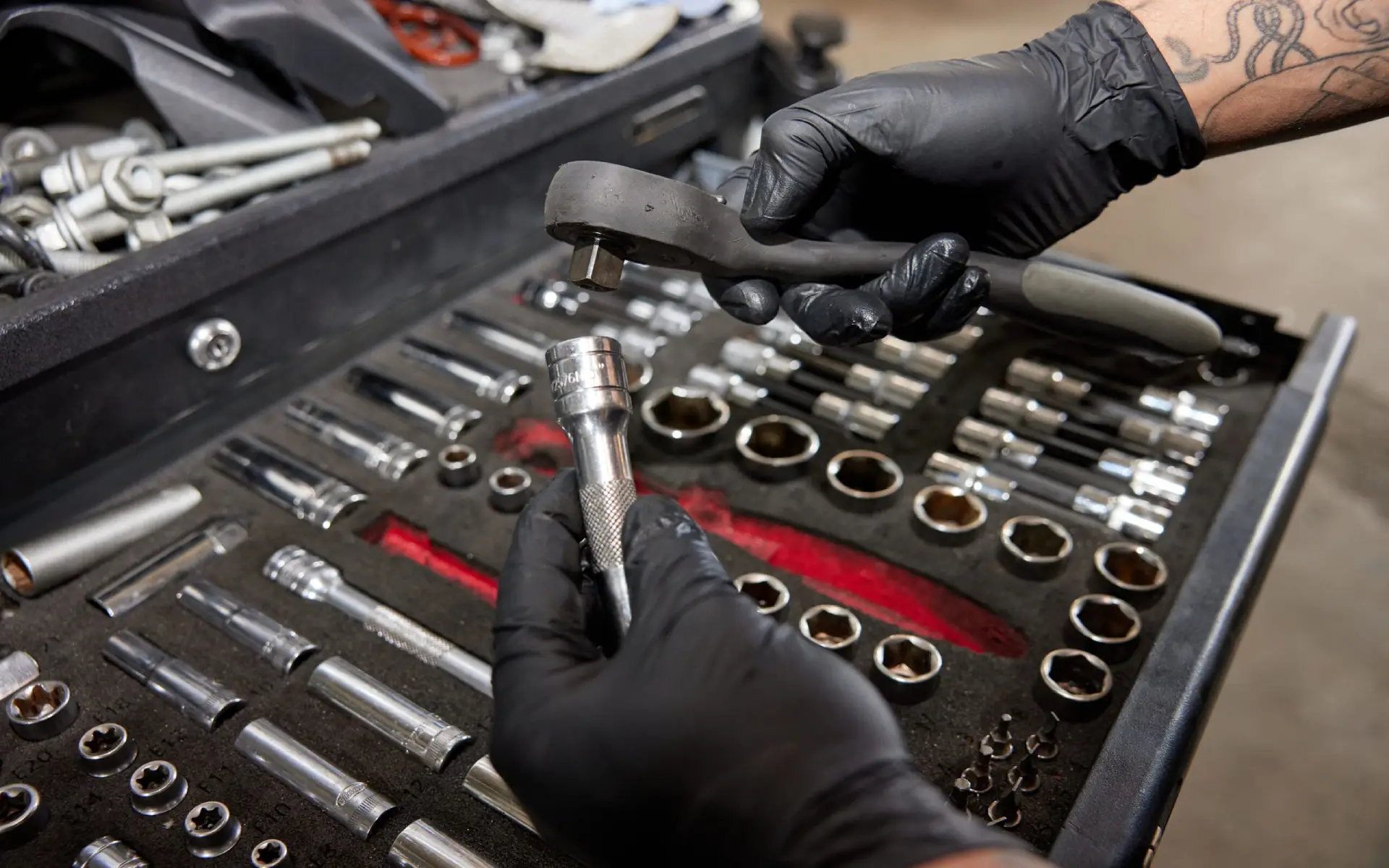 Das Bild zeigt eine Nahaufnahme von Händen in schwarzen Handschuhen, die Werkzeuge aus einem Werkzeugkasten entnehmen. Eine Hand hält eine Ratsche, während die andere einen Steckschlüssel festhält. Im Hintergrund ist eine gut organisierte Sammlung von Stecknüssen und anderen Werkzeugen zu sehen, die ordentlich in einem Schaumstoffeinlagekasten angeordnet sind.